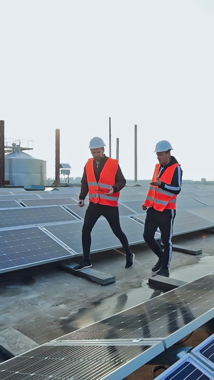 Maintenance in solar power plant. Two male electrician workers in reflective vests and hard hats walking in between long rows of photovoltaic solar panels and talking about installation of new solar panels