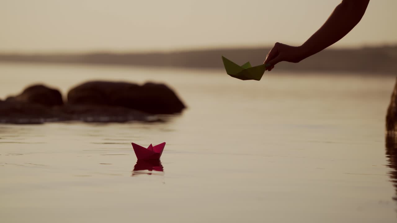 Woman's hand is launching Red and green Paper Ships in the river. Origami Paper Boats are Sailing in the lake. Beautiful Waves Ripple Background. Travel and nature Concept. Filtered video.