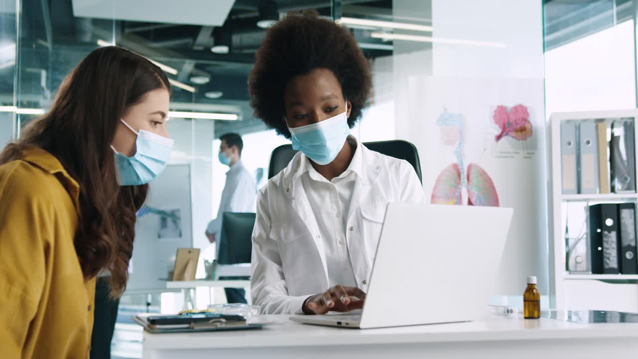 Close-up view of African American female doctor in medical mask using the laptop and explaining to female patient treatment for coronavirus in medical consultation