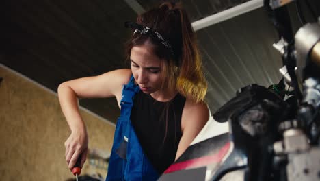 zoom in: a girl auto mechanic repairs a motorcycle and shakes sweat from her forehead with her hand in her workshop. tired of long work in the garage-workshop