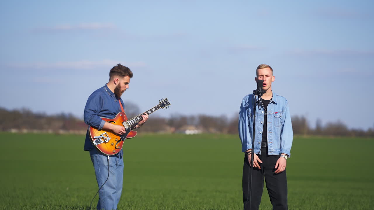 Male singer singing under the guitar music played by bearded musician. Two members of music band perform at the green field backdrop.