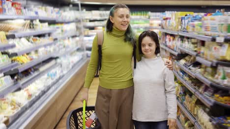 Lächelndes-Mädchen-Mit-Down-Syndrom-Und-Ihrer-Mutter-Im-Supermarkt,-Die-Mit-Einem-Einkaufskorb-Spazieren-Geht