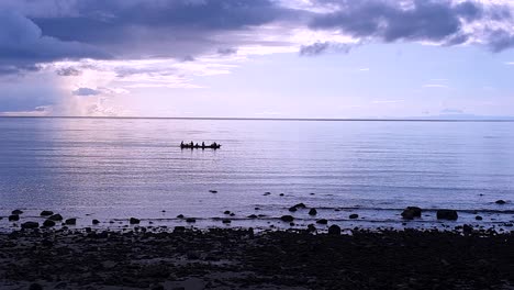 a group of silhouetted local fishermen rowing a traditional wooden fishing boat over the coral reef with beautiful purple haze sunrise on atauro island, timor leste