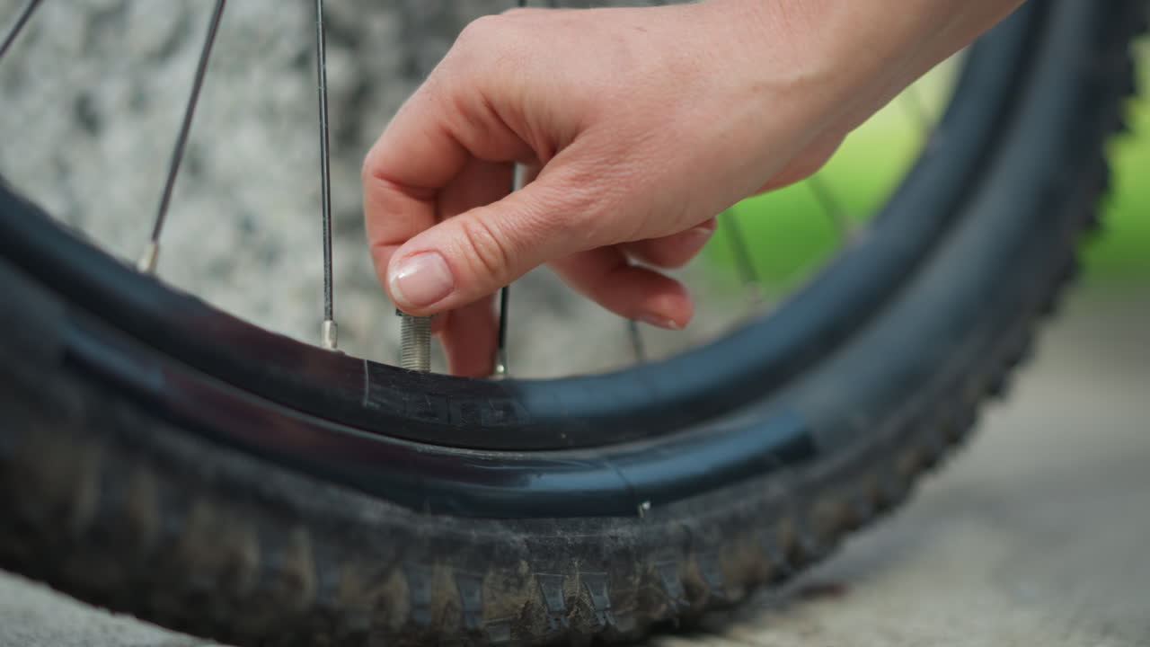 Donna ispeziona la valvola della bicicletta prima di partire, primo piano stretto delle dita che regolano il nucleo della valvola e il tappo sul cerchione sporco, controllo di sicurezza pre-corsa deliberato con accenni di sentiero sullo sfondo sfocato e tattilità attenta