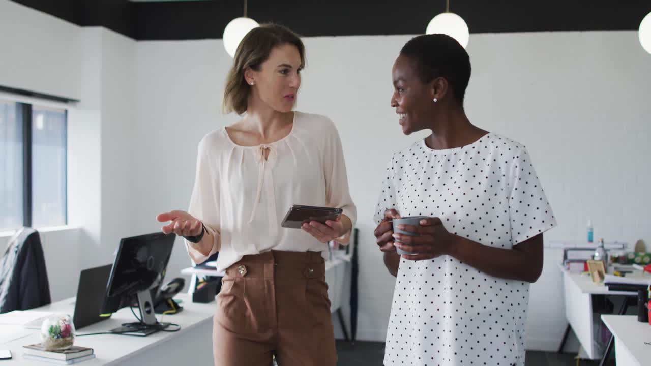 Two diverse female colleagues walking, looking at tablet and discussing in office