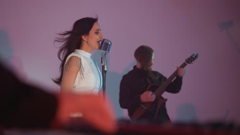 a female singer wearing a white gown stands in front of a vintage microphone, passionately singing. a guitarist plays in the background