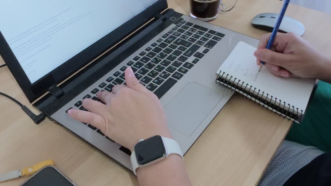 Hands engaged in productive work at a home office desk, writing in a notebook and using a laptop.