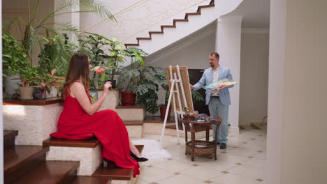 mujer huele a rosa mientras el artista dibuja el retrato de la dama en la mansión. mujer apasionada en vestido rojo con flores posa para el maestro profesional en un apartamento de lujo