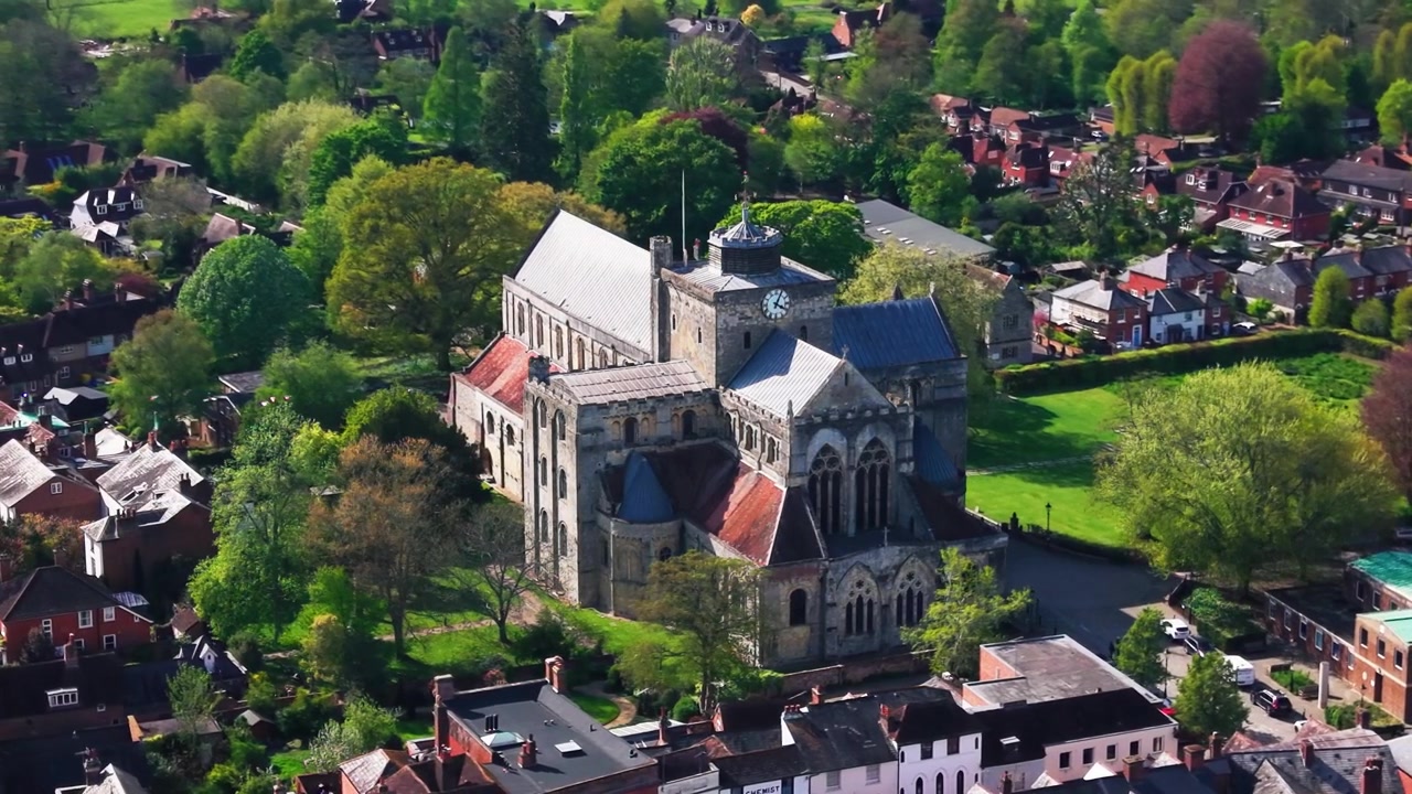 Rekaman drone udara Romsey Abbey di Hampshire, menunjukkan gereja biara bersejarah, arsitektur batu, menara jam, pepohonan di sekitarnya, ruang hijau, atap-atap, dan lanskap kota pada hari yang cerah.