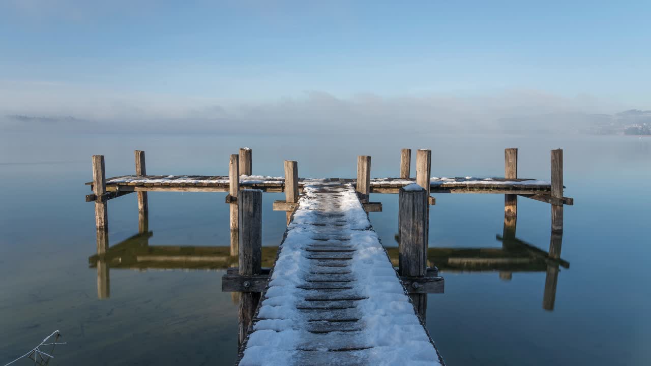 Winter landscape on a lake, on a snow-covered jetty. The fog slowly lifting on the time lapse. The water is calm and there is a calm atmosphere. The sky is blue, the sun is shining