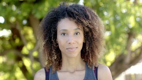 retrato de una feliz mujer biracial con el cabello largo y rizado sonriendo en un jardín soleado, en cámara lenta