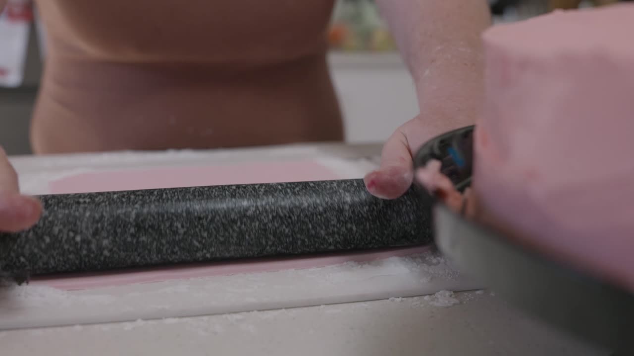 A woman with a brown dress rolls pink fondant icing with a black marbled rolling pin on a white cutting board with a pink cake in the foreground