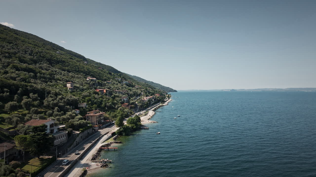 Aproximação cinematográfica de drone 4K UHD voando sobre a exuberante costa de Malcesine, Lago de Garda. Apresenta vilas com telhados vermelhos e água azul — ideal para viagens de luxo e anúncios imobiliários