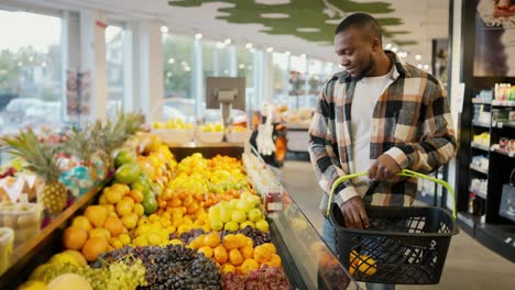 A-guy-with-Black-skin-in-a-checkered-shirt-chooses-citrus-fruits-and-berries-on-the-wide-counter-of-a-modern-supermarket-and-goes-on-with-his-shopping-business