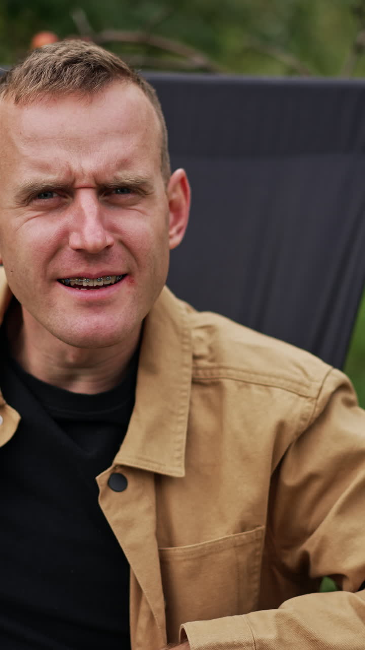 Freelance worker man in a chair looks strictly into camera. Male working remotely in his garden smiling to the camera on his laptop. Blurred nature backdrop. Vertical video