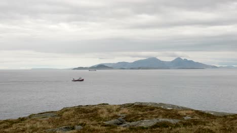 slow panning shot looking out to sea as a red fishing boat moves in front of mountains on scottish islands in the background and heather moorland in the foreground