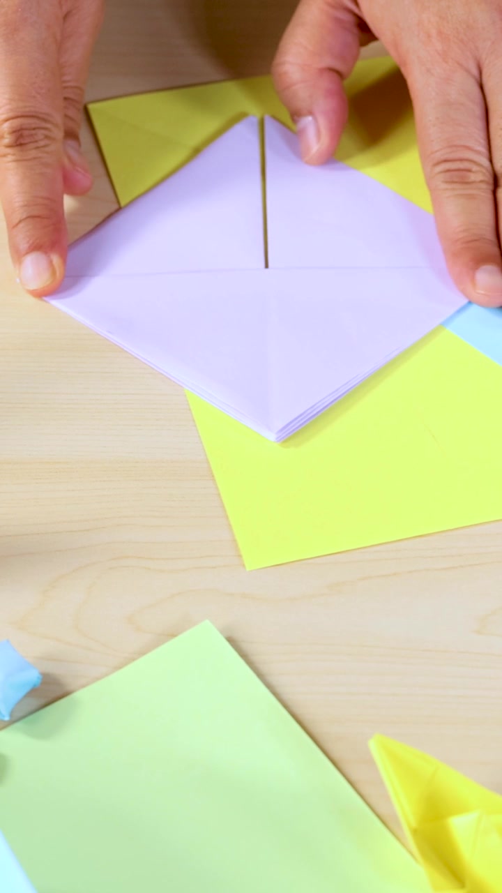 Una persona piega carta pastello su un tavolo di legno, creando origami con un'angolazione della telecamera ravvicinata dall'alto.