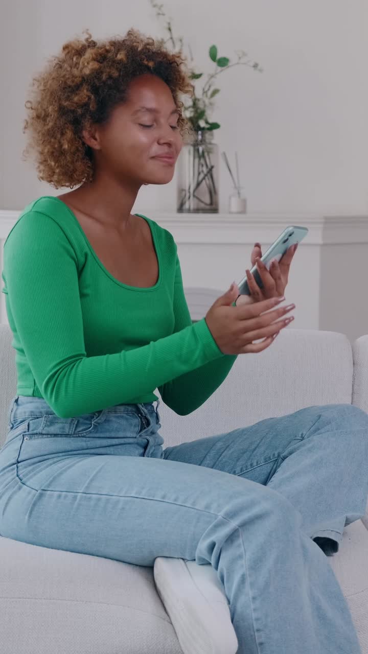 Young woman sits relaxed on a sofa at home scrolling her smartphone and smiling