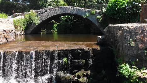 Mit-Einer-Drohne-Aufgenommenes-Video,-Das-über-Den-Wasserfall-Und-Unter-Den-Bögen-Einer-Alten-Brücke-Fliegt