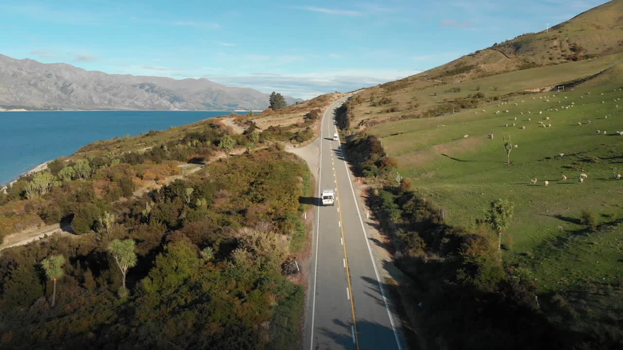 drone aéreo siguiendo una autocaravana con vistas al magnífico lago azul hawea, campo de ovejas y montañas a última hora de la tarde en nueva zelanda