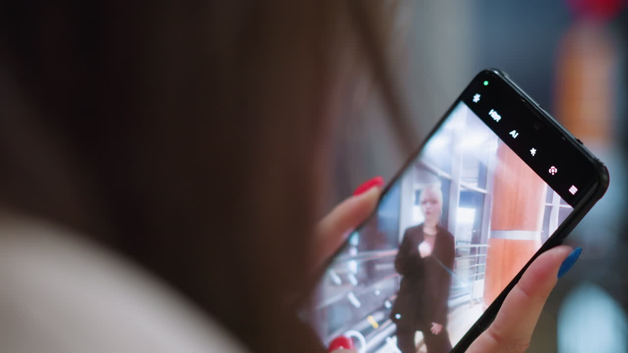 Close up of young lady holding smartphone focused on capturing photo of blonde friend indoors near glass window with city lights reflecting in background