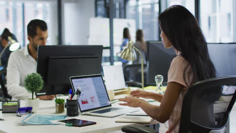 mujer de negocios de raza mixta sentada en un escritorio usando una computadora portátil frente a un colega masculino en una oficina ocupada