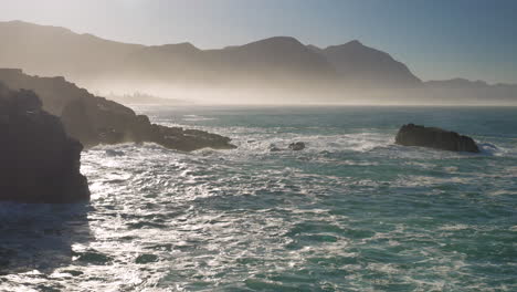 niebla matutina sobre el océano, mientras sale el sol y las olas corren hacia la costa rocosa, hermanus, sudáfrica