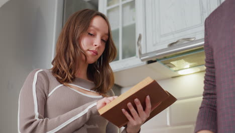 young girl in ribbed sweater reads book thoughtfully, pausing to observe mother cooking in bright kitchen, warm lighting and homey setting create cozy and peaceful atmosphere