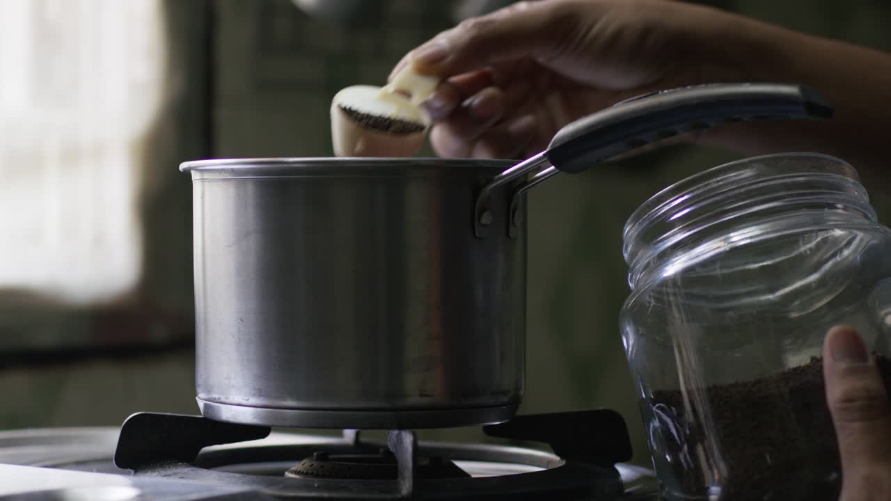 A slow-motion of tea preparation, a hand puts a scoop of tea into a metal pot on the stovetop against a blurred background