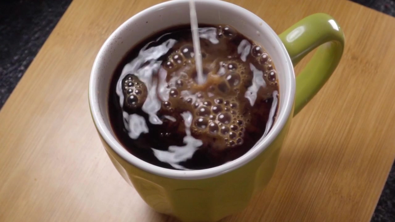Primo piano di latte che si versa nel caffè nero all'interno di una tazza di ceramica. Il liquido che scorre crea motivi a spirale sulla superficie, mescolando colori contrastanti con un movimento circolare fluido.