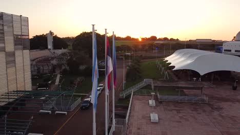 Drone-shot-flying-past-waving-flags-in-front-of-theatre-building-over-the-Parque-del-Conocimiento,-Posadas,-Argentina