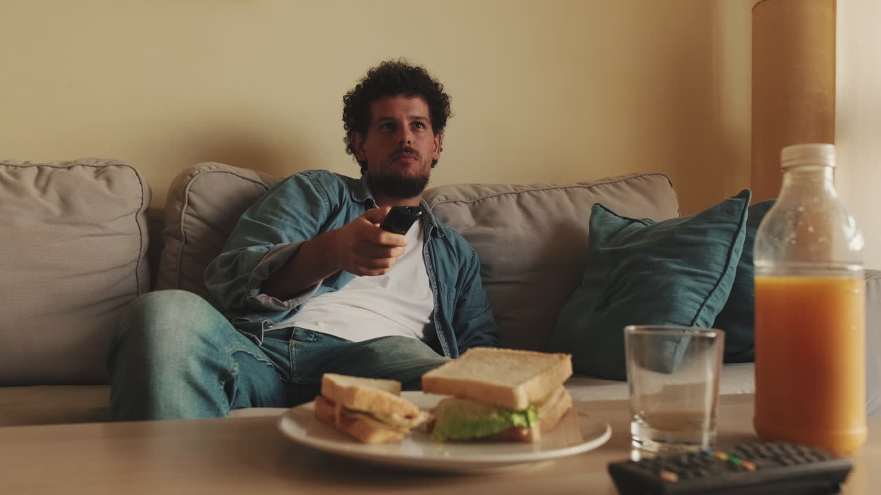 Mand slapper af på sofaen, ser fjernsyn og spiser sandwich