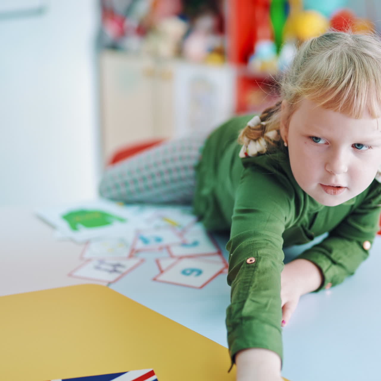 Happy little girl at a table. Curious child sitting alone in the classroom and taking all cards on the table to her place. Cute preschooler with pictures indoors.