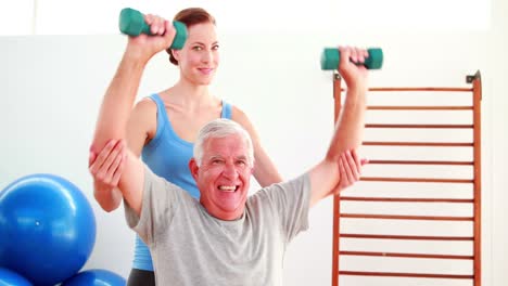 Elderly-man-lifting-hand-weights-sitting-on-exercise-ball-with-his-trainer