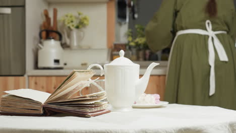 close up of open book on table beside white ceramic mug and glass cup with woman working in background of cozy kitchen, surrounded by flowers, kettle, and calm morning atmosphere