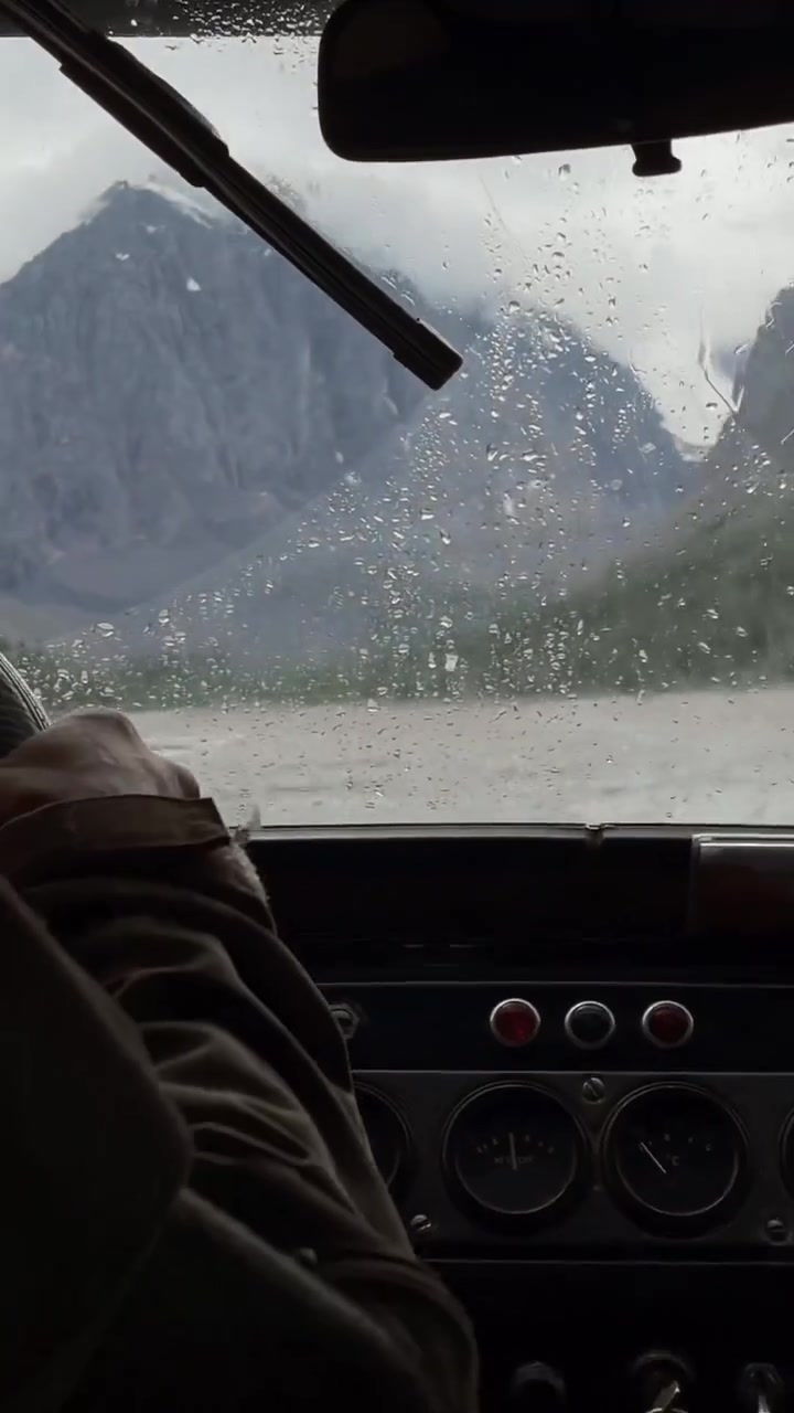 POV dall'interno di un'auto che guida sotto la pioggia, gocce d'acqua sul parabrezza e tergicristallo in movimento, montagne imponenti della Valle di Akturu, Repubblica dell'Altai, Russia.
