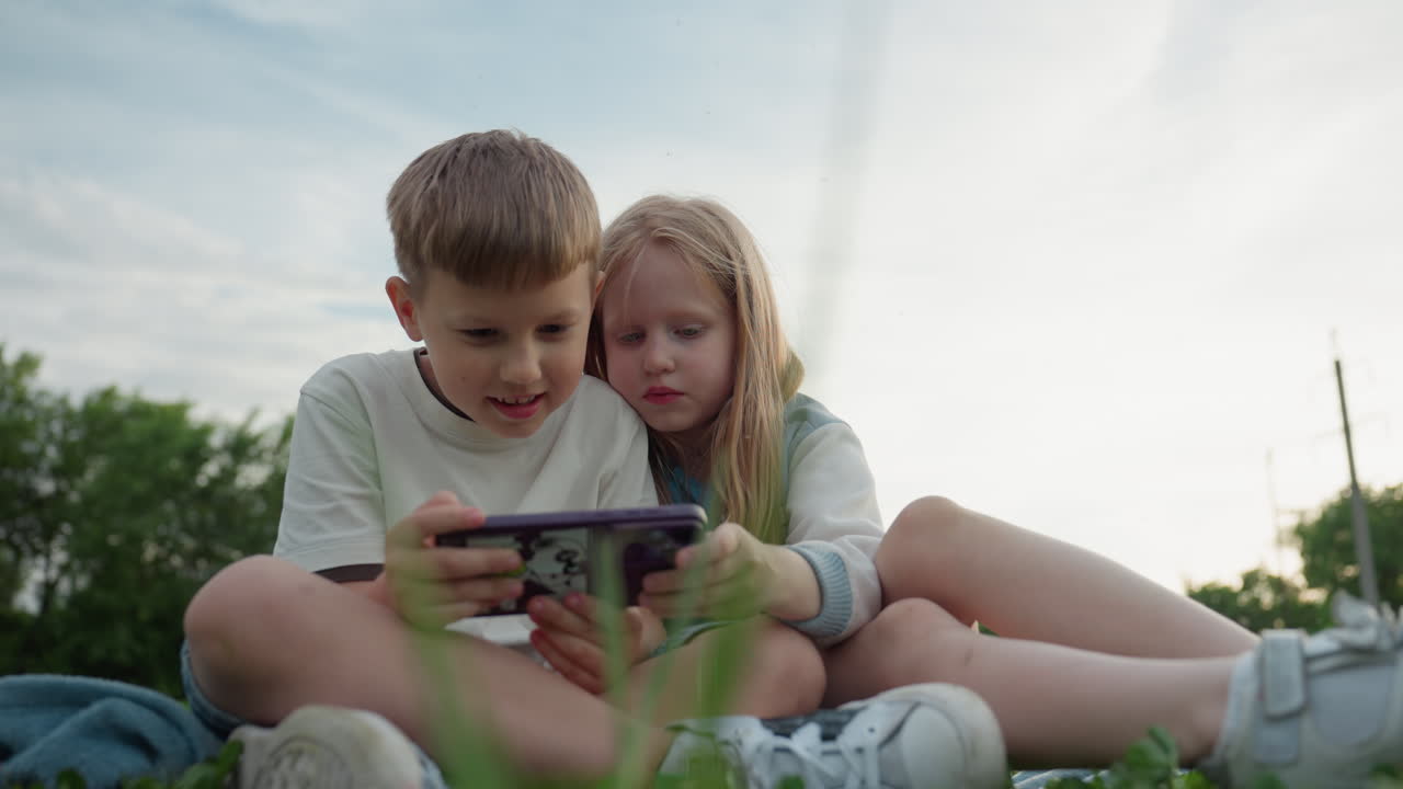 children watching video on phone while sitting on green grass in park, close together with focused expressions, casual summer clothes, blurred trees behind, warm afternoon light creating relaxed mood