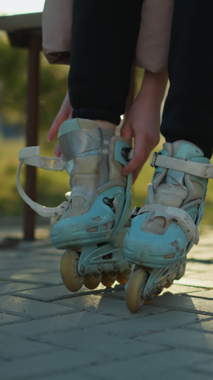 A person wearing a peach jacket and black trousers sits on a park bench, reaching for a rollerblade placed on the ground, preparing to put it on