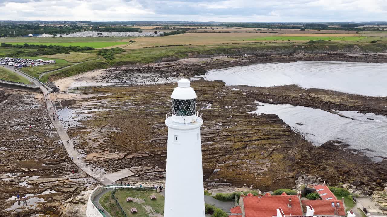 Drone footage circles a white lighthouse overlooking a rocky shoreline and tidal pools under cloudy daylight, revealing coastal landscape and nearby buildings