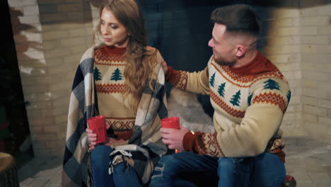 young couple at christmas time at home. man and woman in christmas sweaters sitting near fireplace with red cups. romantic atmosphere of couple in love.