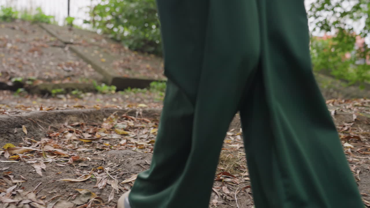 Donna bianca che percorre un sentiero alberato in un parco con passo sicuro e passo cauto su radici esposte, pendolare trasformatasi in esploratrice che si muove su un terreno umido, scarpe da ginnastica e pantaloni verdi visibili, pausa per riprendere fiato