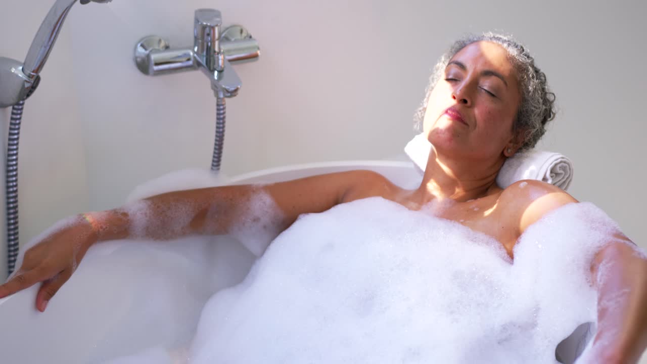 Vertical video: Sunlight shifting, woman reclining and adjusting arm in bathtub with foam for rest