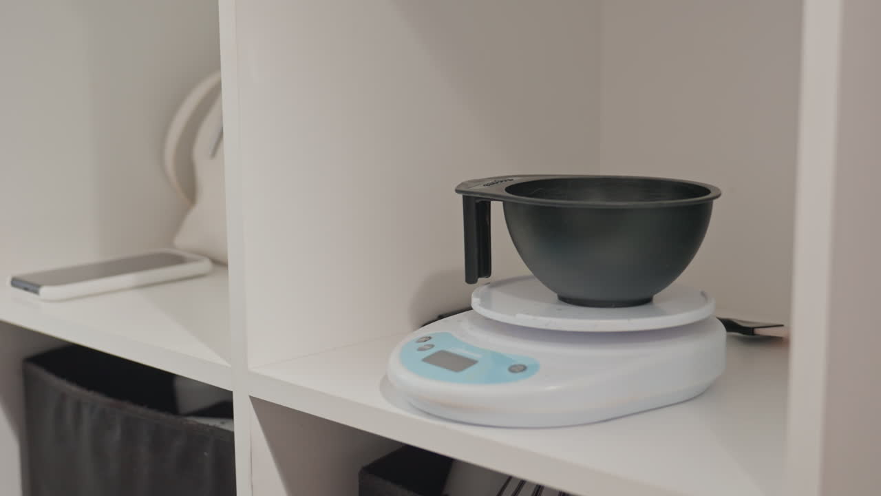 Salon mixing bowl on scale measuring powder for precise hair color prep gloved hand adds powdered pigment into black bowl on digital scale inside white shelving station, clinical clean aesthetic
