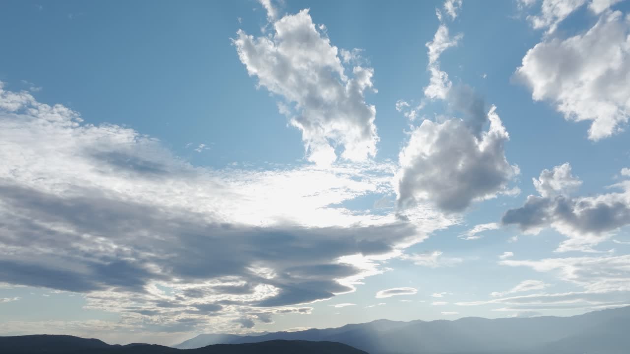 Beautiful afternoon drone shot in 70mm showing pale mountains and a blue sky filled with white clouds, some shining brightly from sunlight. The sun is hidden behind the clouds.