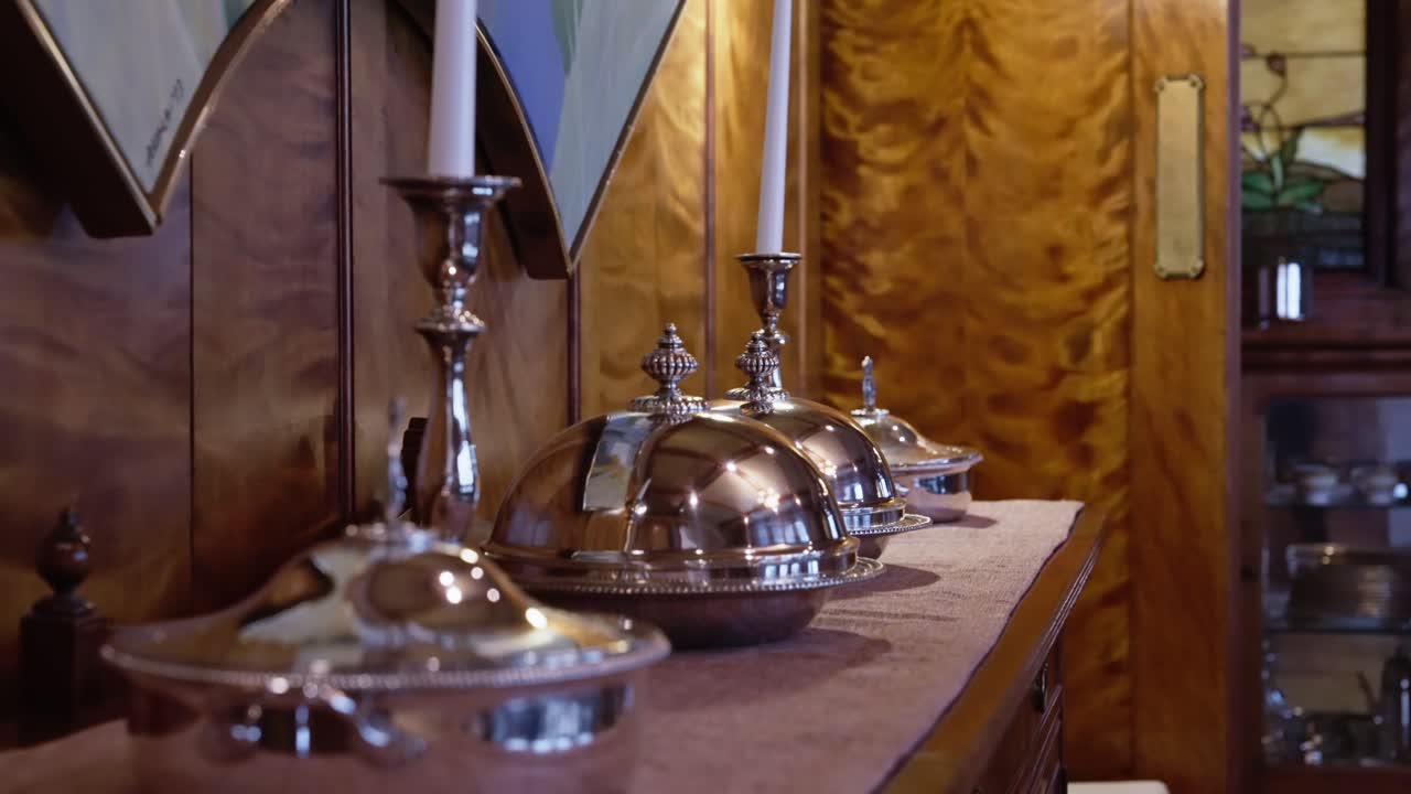 Silver serving dishes and tall candlesticks on a wood buffet in an ornate dining room