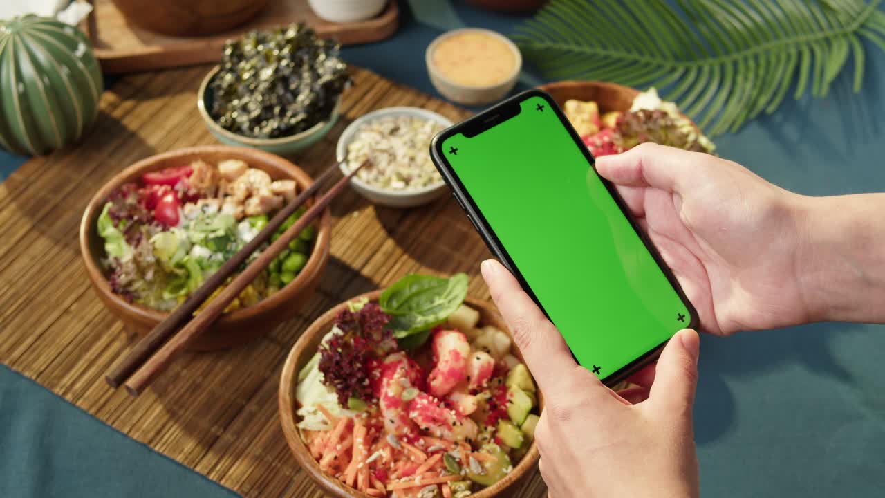 tazones de poke hawaianos, sosteniendo un teléfono inteligente con pantalla verde cromada de primer plano. poke cocido hecho de verduras en rodajas, mariscos y verdura. platos vegetarianos saludables. comida cruda vegana asiática, palillos
