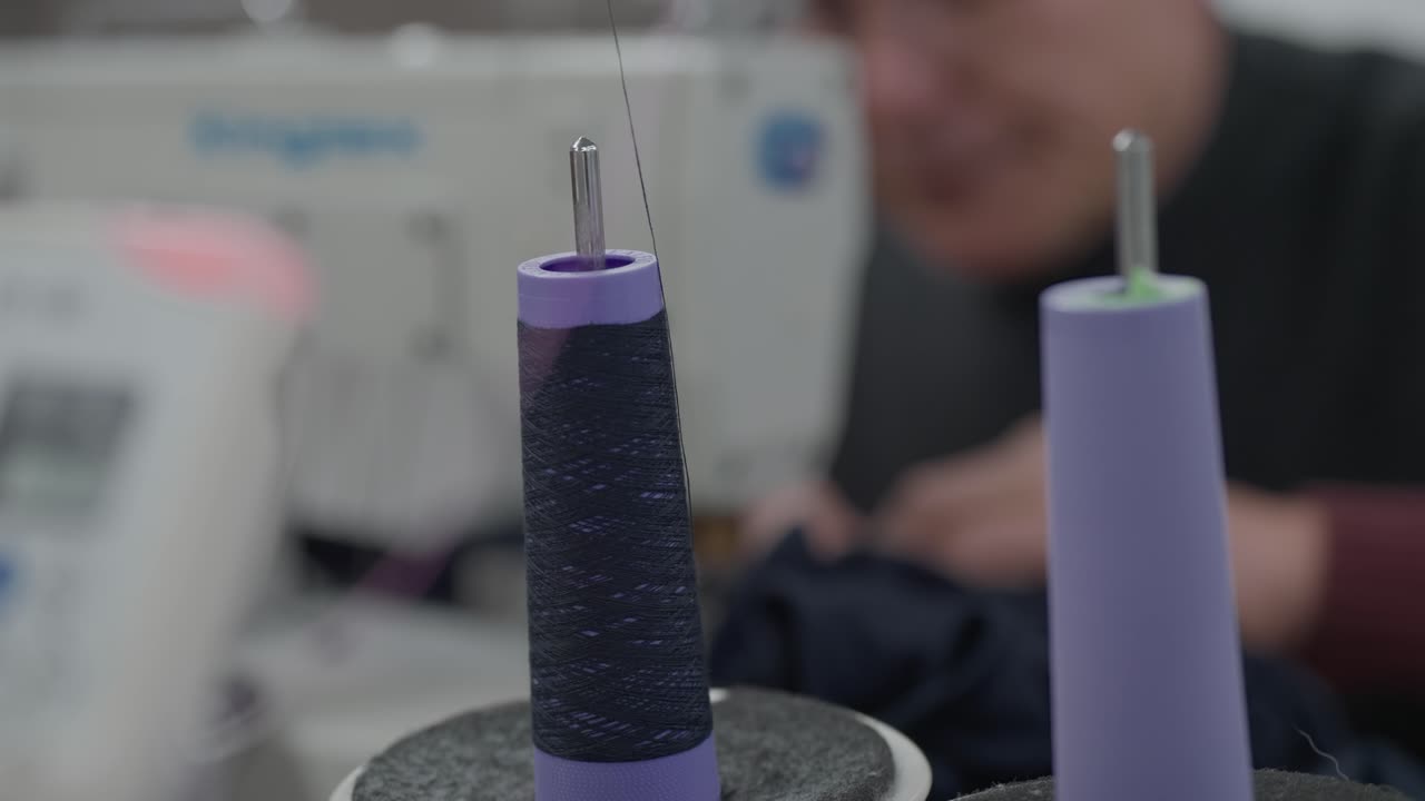 Two dark navy thread cones on sewing machine spindle pins with blurred worker in background
