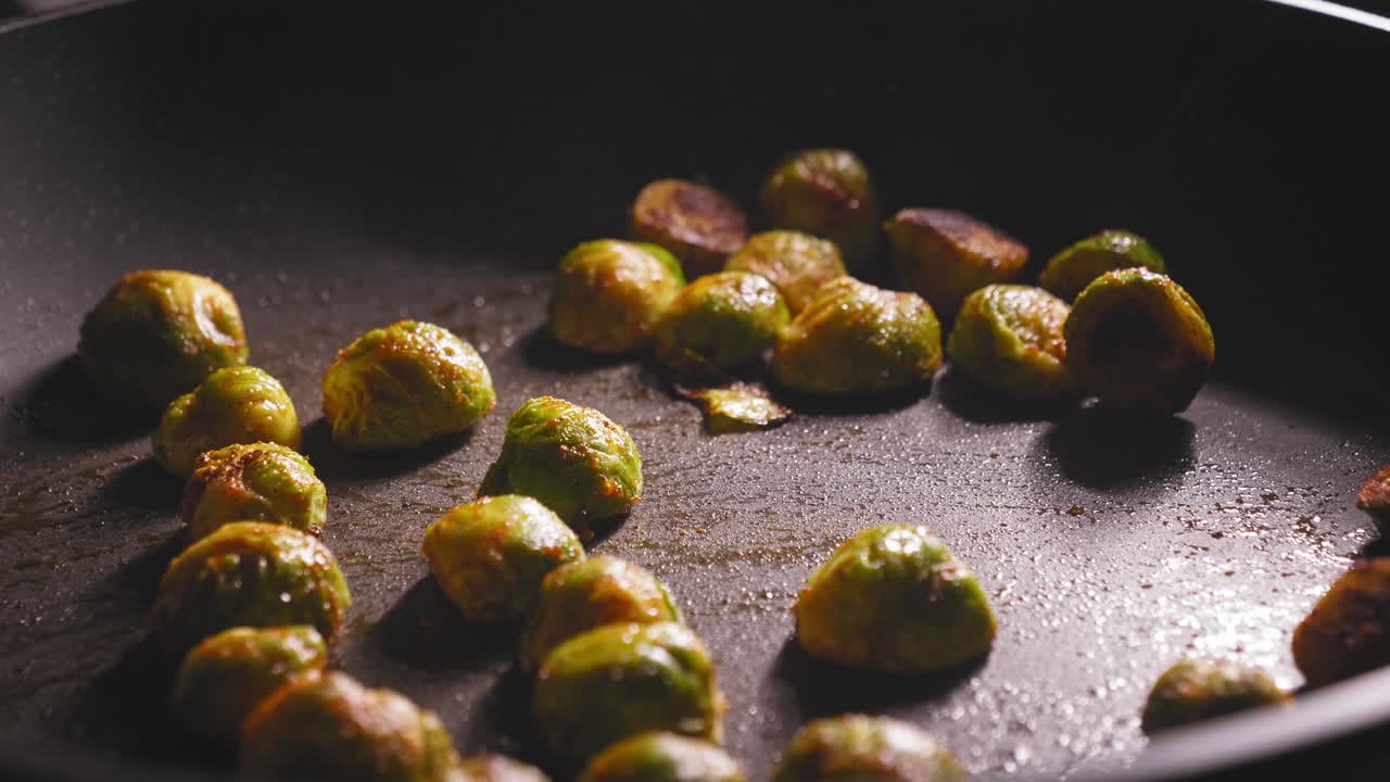 A close up shot of marinated brussels sprouts roasting in a hot frying pan, a chef holding a spatula carefully turns the sprouts with a wooden spatula to ensure an even cook