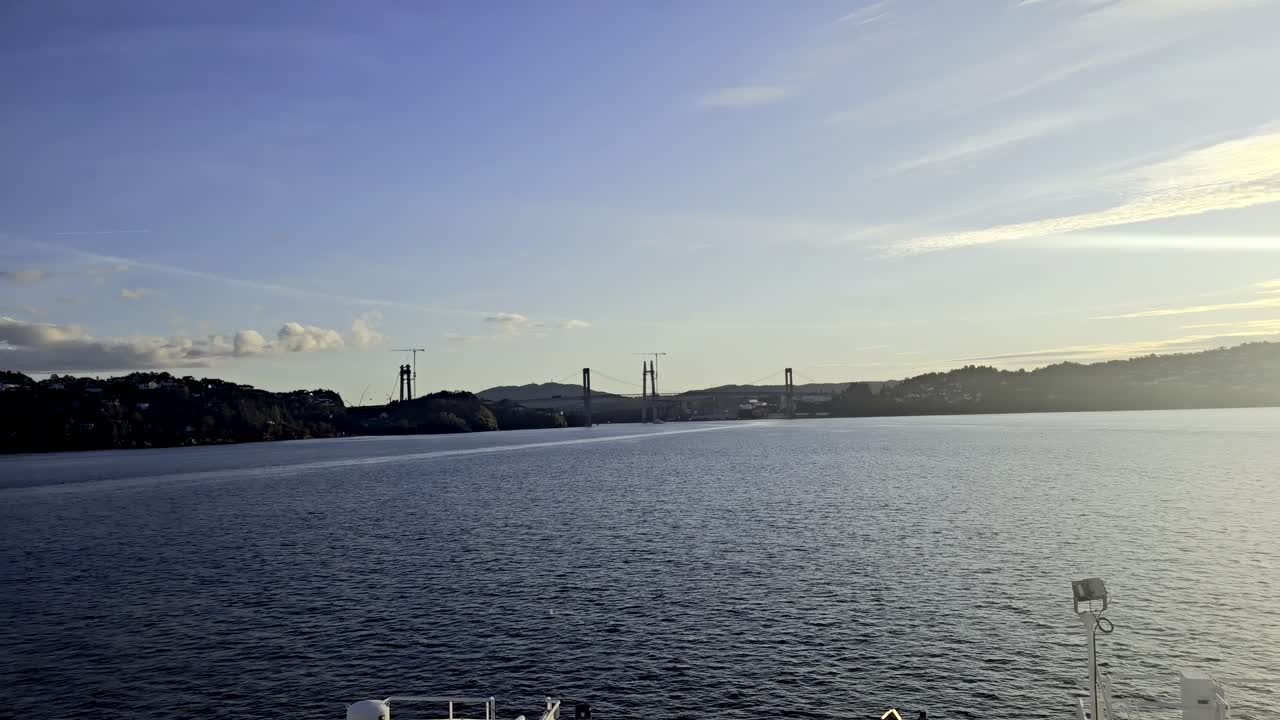 Wide view from vessel showing open sea and distant Sotra Bridge towers with cranes in golden evening light