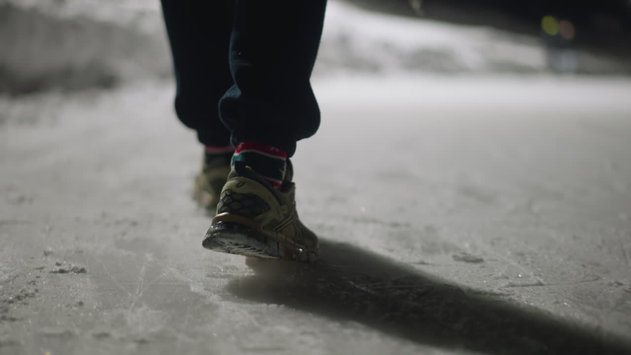 Rear leg view of individual walking on icy surface at night while holding object in hand, wearing sneakers and festive socks, with long shadow cast on frozen ground under dim winter lighting